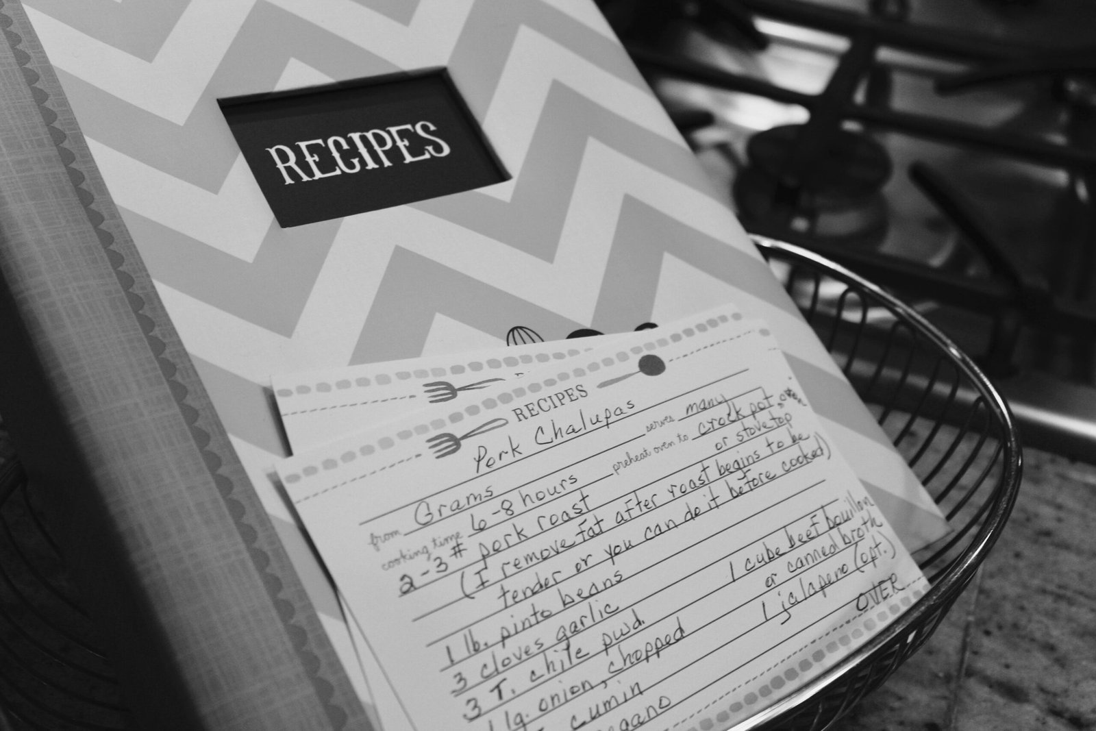 Monochrome image of a recipe book and handwritten notes in a kitchen setting.